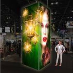 Trade show booth: green panel with glowing lanterns and a woman's portrait; a presenter stands beside it.
