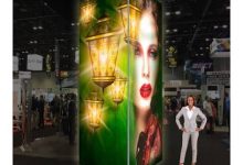 Trade show booth: green panel with glowing lanterns and a woman's portrait; a presenter stands beside it.