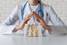 Doctor with stethoscope protecting a paper-family and wooden house, symbolizing family health insurance/care