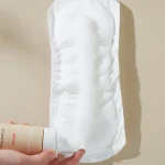 Hands hold a white disposable sanitary pad/diaper insert against a beige background, with a lotion bottle visible in the lower left corner