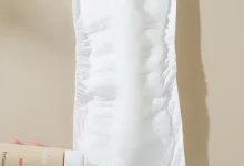 Hands hold a white disposable sanitary pad/diaper insert against a beige background, with a lotion bottle visible in the lower left corner