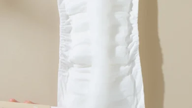 Hands hold a white disposable sanitary pad/diaper insert against a beige background, with a lotion bottle visible in the lower left corner