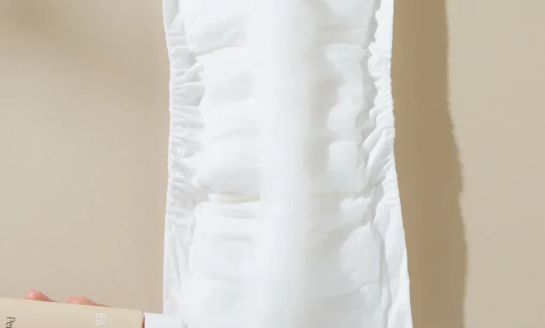 Hands hold a white disposable sanitary pad/diaper insert against a beige background, with a lotion bottle visible in the lower left corner