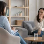 Two women in a therapy session seated on a sofa, one taking notes on a clipboard while the other listens thoughtfully.