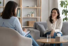 Two women in a therapy session seated on a sofa, one taking notes on a clipboard while the other listens thoughtfully.