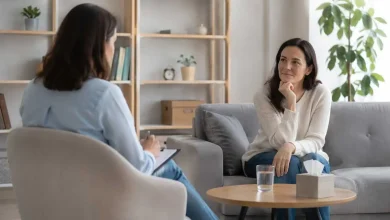 Two women in a therapy session seated on a sofa, one taking notes on a clipboard while the other listens thoughtfully.